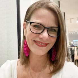 Woman smiling confidently with stylish accessories. A smiling woman with glasses and pink earrings in a white top, standing indoors.