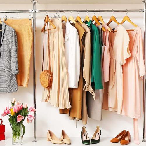 Elegant Wardrobe Styling with Neutral and Pastel Outfits and Accessories A stylish and organized clothing rack featuring a collection of neutral and pastel-colored outfits, including blazers, dresses, and tops in beige, white, green, and blush tones. Accessories include a woven crossbody bag and various pairs of heels in nude and green shades. A bouquet of pink tulips and a red handbag sit on the floor beside the rack, adding a fresh and vibrant touch.