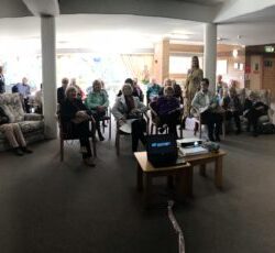 Community Event – Engaging Group Discussion in a Relaxed Setting A group of elderly individuals seated in a semi-circle in a spacious community room with brick walls, a fireplace, bookshelves, and large windows letting in natural light. A presentation setup with a laptop and projector is visible at the center.