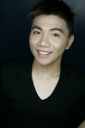 Professional Headshot – Young Man with Modern Hairstyle and Friendly Smile A professional headshot of a young man with short, dark hair styled upwards. He has a bright smile, smooth complexion, and is wearing a black V-neck T-shirt against a dark background.