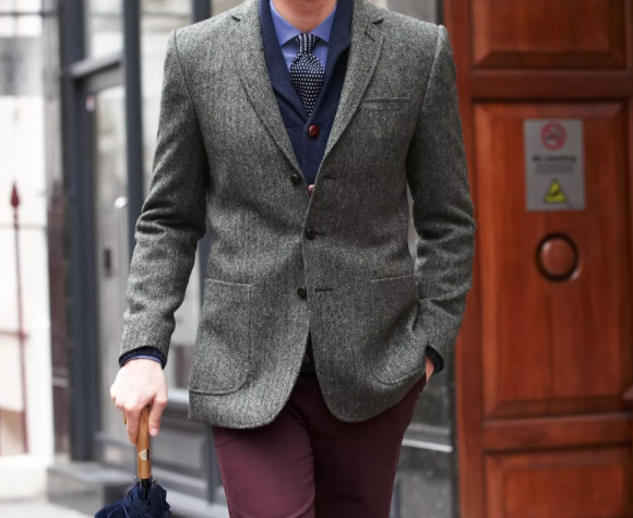 How Men's Fashion Works – Classic Tweed Blazer & Smart Casual Styling A stylish man walking down a city street wearing a tailored tweed blazer over a navy cardigan, blue shirt, and patterned tie. He pairs the outfit with burgundy trousers and carries a closed umbrella.