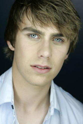 Professional Headshot – Young Man with Blue Eyes and Casual Style A professional headshot of a young man with tousled brown hair, blue eyes, and a confident expression. He is wearing a white button-up shirt against a dark background.