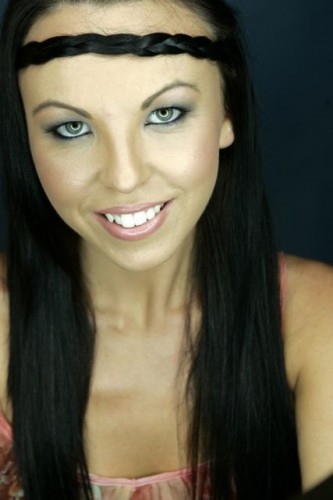Professional Headshot – Young Woman with Braided Hairband and Green Eyes A professional headshot of a young woman with long, straight black hair styled with a braided headband. She has striking green eyes, a warm smile, and is wearing a floral top against a dark background.