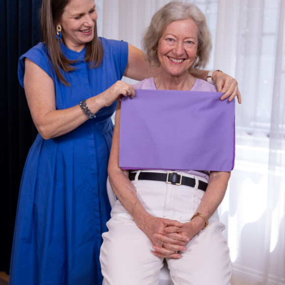 Older woman smiling as a personal stylist holds up a lilac drape for colour matching