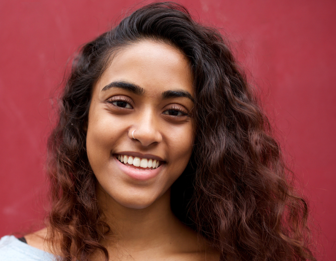 Hairstyle Session – Elegant Braided Updo in Progress Young woman with long curly hair and nose ring, smiling warmly against a deep red background.