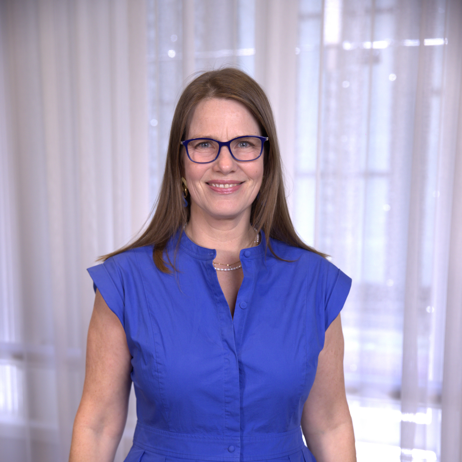 Woman in blue glasses and dress smiling in front of sheer curtains