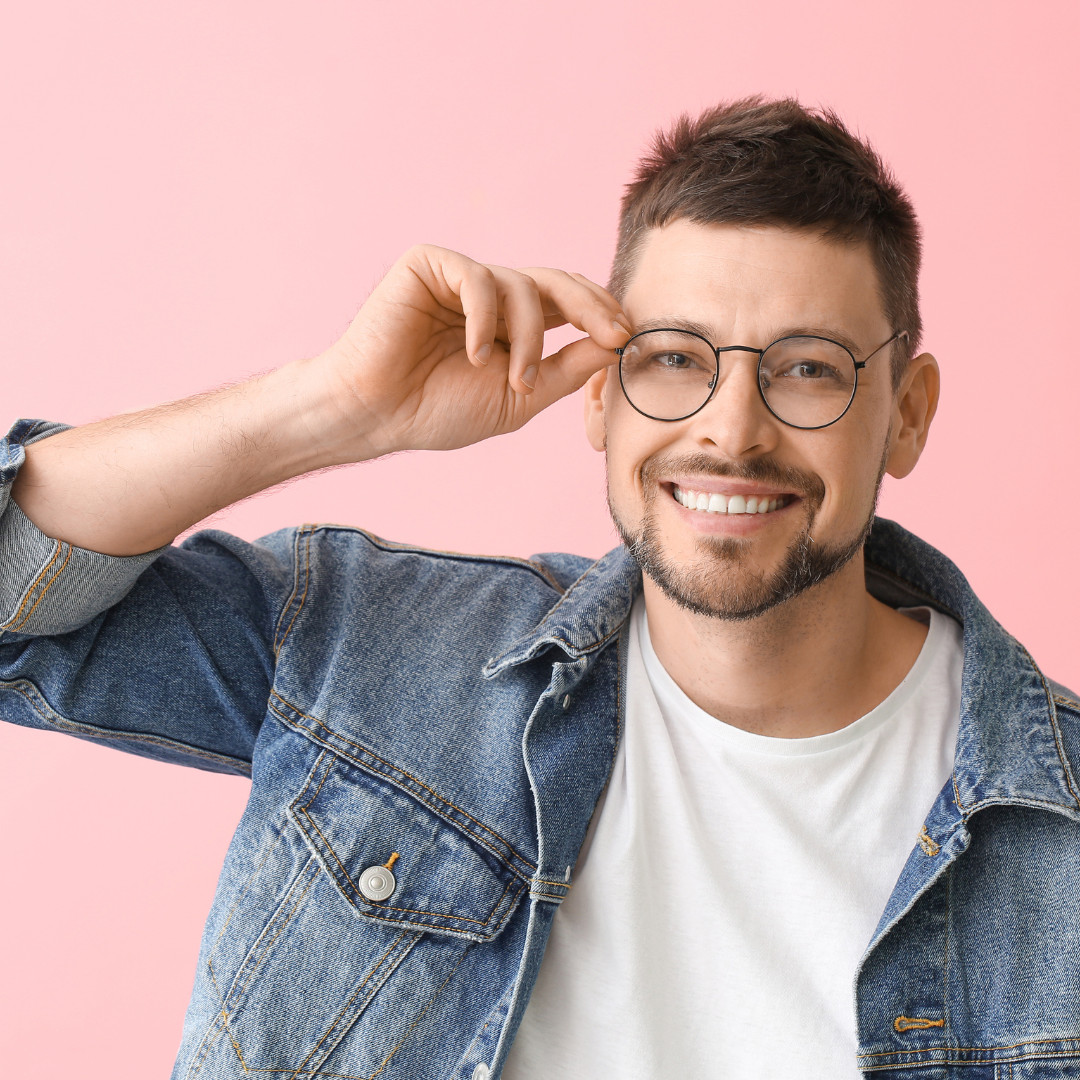 Virtual Eyewear Style Session – Trendy Round Glasses with Bold Frame Design Young man with glasses smiling, wearing a denim jacket and white t-shirt, posing against a soft pink background.